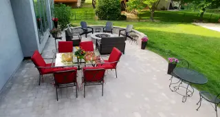 Large paver patio with red-cushioned dining set, gas fire pit surrounded by lounge chairs, and bistro table overlooking a green lawn